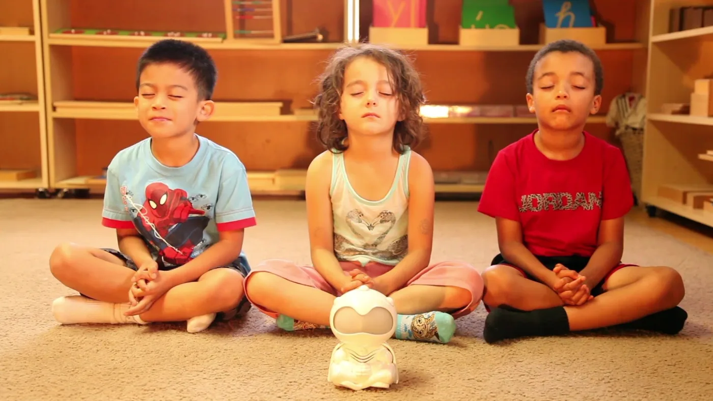 Three kids meditating cross-legged with an early GLY device guiding them during a school session